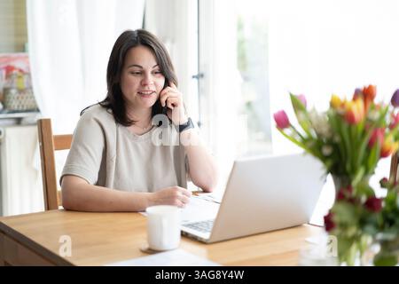Belle fille brune souriante assise à table devant un ordinateur portable et des documents parlant au téléphone. Concept de bureau à domicile Banque D'Images