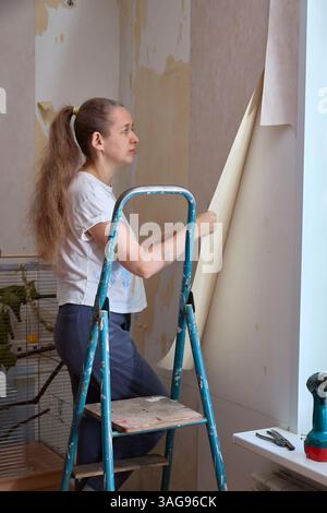 Femme sur l'échelle enlevant soigneusement le vieux papier peint dans une pièce, révélant des couches en dessous. Elle se concentre sur la tâche, engagée dans un projet de rénovation domiciliaire Banque D'Images