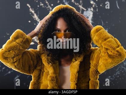 Femme à la mode en manteau de fourrure jaune ajustant la capuche, portant des lunettes de soleil élégantes. style, tendance, vêtements d'extérieur, hiver, streetwear, accessoires Banque D'Images