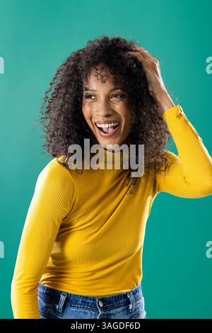 Femme souriante en pull jaune touchant les cheveux bouclés sur fond vert. Portrait, mode, joyeux, confiant, coiffure, dynamique Banque D'Images