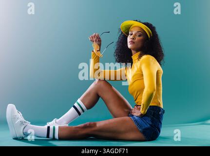 Femme confiante en haut jaune et visière assis, tenant des lunettes de soleil. Mode, style, accessoires, tendance, été, décontracté Banque D'Images