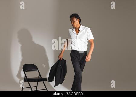 Homme confiant dans la tenue élégante marchant devant la chaise dans le studio moderne. Mode, style, confiance, urbain, contemporain, chic Banque D'Images