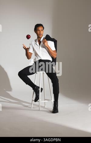 Homme élégant en chemise blanche jonglant avec la pomme tout en étant assis sur le tabouret dans le studio. Mode, masculin, style de vie, contemporain, équilibre, loisirs Banque D'Images