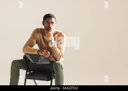 Homme afro-américain dans la tenue élégante assis sur la chaise, regardant soigneusement devant. Mode, contemplation, moderne, élégance, style de vie Banque D'Images