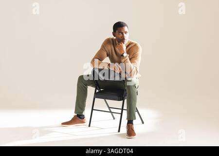 Homme afro-américain dans une tenue élégante assise sur une chaise, regardant réfléchi. Mode, style, pensif, contemplation, élégance, portrait Banque D'Images