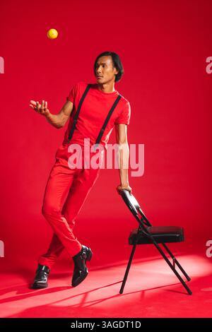 Homme dans la tenue rouge équilibre sur la chaise, lançant la balle jaune dans le studio. Jonglerie, équilibre, coordination, performance, athlétique, acrobaties Banque D'Images