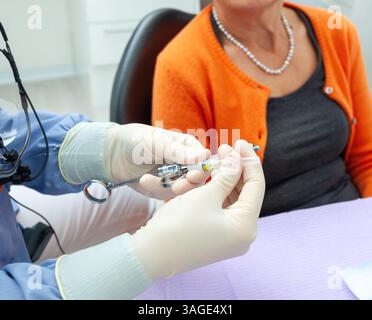 Dentiste portant une loupe et des gants chirurgicaux préparant une seringue anesthésique pour patient senior en clinique dentaire Banque D'Images