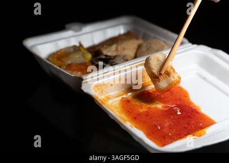 Main tenant des baguettes avec un morceau de nourriture plongé dans un récipient de sauce. Les aliments sont placés dans une boîte à emporter blanche avec divers plats v Banque D'Images