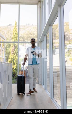 Afro-américain homme afro-américain marchant avec valise et smartphone dans le couloir moderne. Affaires, voyageur, technologie, transport, entrepreneur, Banque D'Images