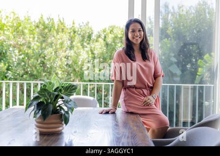 Femme souriante en robe rose assise sur la table près de la grande fenêtre à la maison. Style de vie, mode, féminin, élégance, loisirs, relaxation Banque D'Images