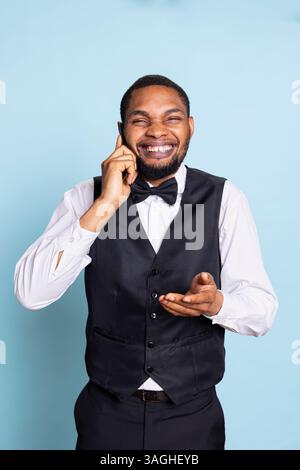 Chasseur afro-américain effectuant des réservations de chambres par appel téléphonique, partageant des informations sur les équipements de l'hôtel sur fond bleu. Le jeune portier répond aux questions en studio. Banque D'Images