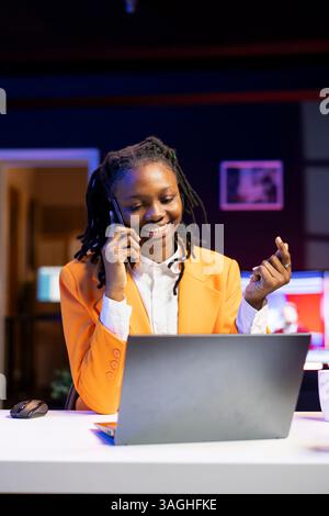 Femme heureuse recevant un appel téléphonique agréable d'un ami tout en travaillant à la maison sur ordinateur portable dans le bureau personnel. Gai freelance parlant avec mate en ligne à l'aide du téléphone portable tout en résolvant des tâches Banque D'Images