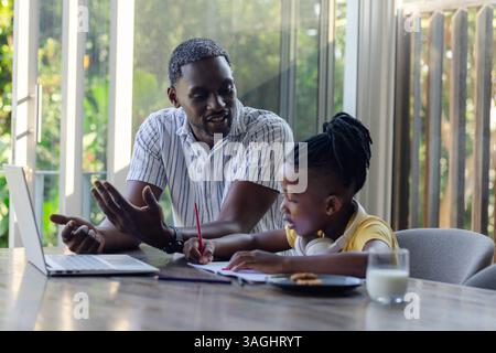 Père afro-américain aidant son fils à faire ses devoirs à la maison, en utilisant un ordinateur portable et en écrivant des notes. Parentalité, tutorat, éducation, technologie, mentorat, goujon Banque D'Images