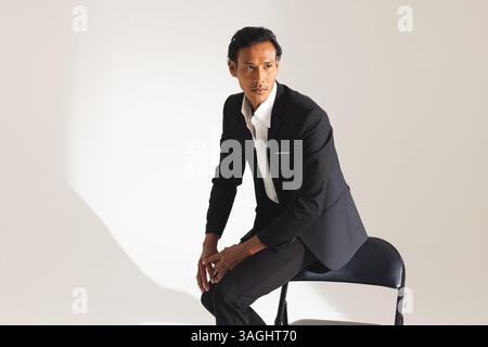 Homme confiant en costume noir assis sur la chaise, regardant soigneusement loin. Affaires, professionnel, exécutif, mode, formel, contemplatif Banque D'Images