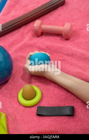 Gros plan d'une personne pressant une balle bleue pour la thérapie de la main, entourée d'outils d'exercice sur une serviette rose. Banque D'Images