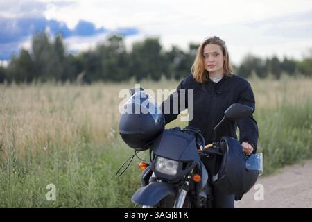 Une femme debout à côté d'une moto dans un paysage rural, portant une veste noire et tenant un casque Banque D'Images