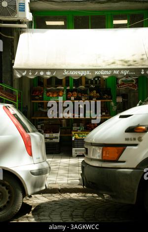 Petite boutique de légumes en bordure de rue avec façade peinte en vert, partiellement cachée derrière des voitures garées, dans une zone urbaine animée. Banque D'Images