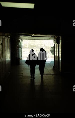 Silhouettes de deux personnes marchant à travers un passage souterrain piétonnier faiblement éclairé vers la lumière du jour, scène urbaine tunnel. Banque D'Images