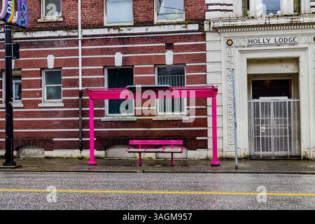Abri d'arrêt de bus rose avec un banc devant un bâtiment en briques. Surface de rue humide, lumière nuageuse du jour, et aucune personne présente Banque D'Images