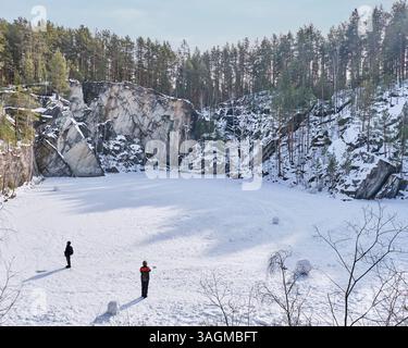 Sysert, Russie - 5 mars 2025 : deux personnes se tiennent debout sur un lac gelé recouvert de neige dans une carrière de talc entourée de falaises rocheuses et d'une forêt de pins. Sceni Banque D'Images