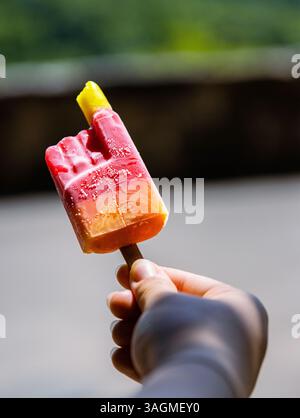Gros plan d'une crème glacée rouge jaune orange mordue colorée avec bâton de popsicle tenu dans une main d'enfant méconnaissable. Journée d'été ensoleillée dans le parc Banque D'Images