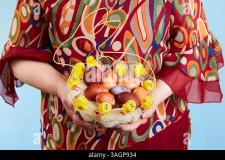 Une grand-mère tenant un petit panier tissé en forme de nid rempli d'œufs de Pâques décorés et de petites figurines de poussin jaunes. Banque D'Images