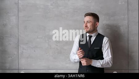 homme en vêtements d'affaires se tient contre le mur, ajustant les poignets de sa chemise, avec un look concentré. L'arrière-plan neutre et l'éclairage doux créent un atm Banque D'Images