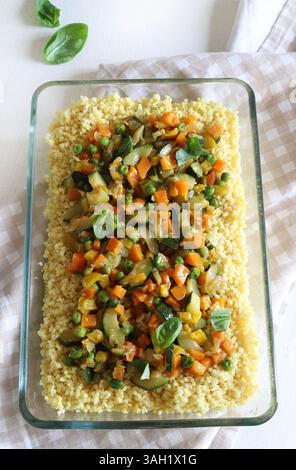 Couscous aux légumes (courgettes, poivrons, carottes, herbes) dans un plateau en verre isolé sur une table en bois blanc. Nourriture saine et végétarienne. Banque D'Images
