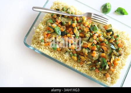 Couscous aux légumes (courgettes, poivrons, carottes, herbes) dans un plateau en verre isolé sur une table en bois blanc. Nourriture saine et végétarienne. Banque D'Images
