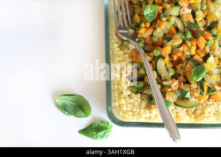 Couscous aux légumes (courgettes, poivrons, carottes, herbes) dans un plateau en verre isolé sur une table en bois blanc. Nourriture saine et végétarienne. Banque D'Images