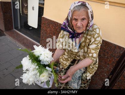 LVIV, UKRAINE - 20 juin 2021 : une femme âgée est assise dans la rue, tenant un bouquet de fleurs blanches tout en les vendant aux passants. Sa tenue traditionnelle reflète la culture et l'histoire locales. Banque D'Images