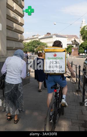 LVIV, UKRAINE - 20 juin 2021 : un cycliste de livraison de nourriture passe devant des piétons dans une rue animée, portant un sac de livraison sur son dos tout en présentant un service alimentaire local. Banque D'Images