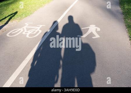 Un couple marchant côte à côte projetant de longues ombres dans le soleil de fin d'après-midi sur un sentier / piste cyclable séparé à Eastrop Park, Basingstoke, Royaume-Uni Banque D'Images