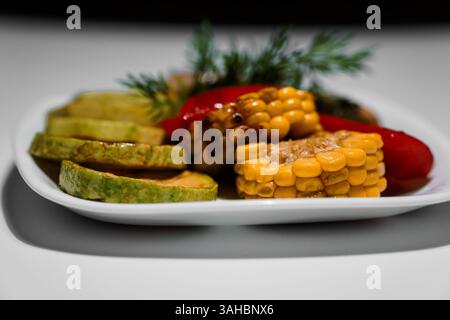 Gros plan d'une assiette blanche avec du maïs grillé sur l'épi, des tranches de courgettes et des poivrons rouges garnis de brins d'aneth frais. Banque D'Images