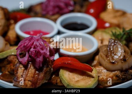 Délicieux plateau de viande grillée avec légumes colorés, sauces, oignons marinés et avocat servis sur plaque blanche vue en gros plan Banque D'Images
