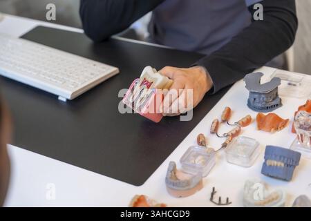 Dentiste expliquant l'anatomie dentaire à l'aide d'un modèle lors d'une consultation, entouré de divers prothèses et outils dentaires Banque D'Images