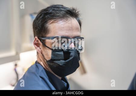 Dentiste portant des loupes chirurgicales et un masque effectue habilement des procédures dentaires dans une clinique dentaire moderne, assurant les soins et l'hygiène des patients Banque D'Images