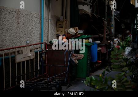 Le collecteur d'ordures prend une pause à côté de son chariot et de ses poubelles dans une ruelle cachée de Bangkok Banque D'Images
