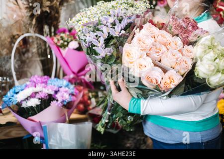 Fleuriste tenant plusieurs bouquets enveloppés, y compris des roses, des lis et des marguerites dans un studio de fleurs animé. Arrangements faits main, floraison saisonnière Banque D'Images