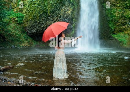 Femme tenant un parapluie rouge se tient dans l'eau peu profonde près d'une cascade, entourée d'une végétation luxuriante. Silver Falls, Oregon, États-Unis Banque D'Images