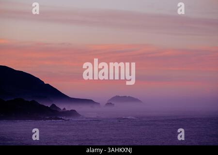 Vue sur l'océan sereine au coucher du soleil avec des montagnes brumeuses à l'horizon et un ciel rose-orange. Monterey, CALIFORNIE, États-Unis Banque D'Images