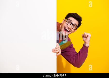 Jeune homme joyeux avec des lunettes célébrant et jetant un œil derrière un panneau blanc sur un fond jaune vif Banque D'Images