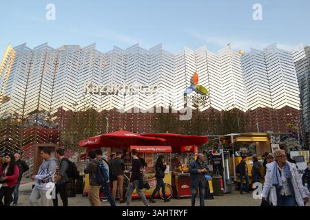 La façade du pavillon du Kazakhstan avec des panneaux de couleur argentée d'isolation thermique reflète le pavillon des Émirats arabes Unis sur fond de ciel bleu. Petit marché sur la place. Banque D'Images