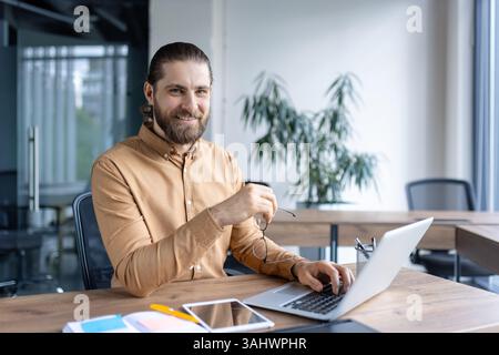 Un homme d'affaires souriant à son bureau, tenant des lunettes, travaille sur son ordinateur portable dans un cadre de bureau moderne. L'homme regarde directement la caméra, rayonnant de confiance. Banque D'Images