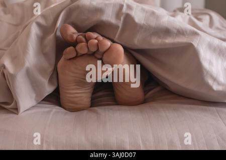 Gros plan des pieds d'une personne dormant au lit. Semelle des pieds masculins dépassant de la couette dans le lit faible profondeur du champ vue avant. Plan d'un homme pieds Banque D'Images