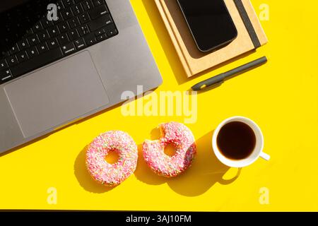 Espace de travail avec ordinateur portable, café et beignets sur fond jaune – Flat lay Banque D'Images