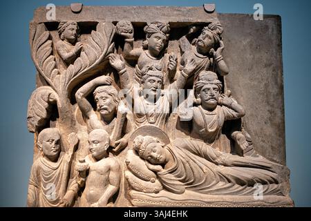 WASHINGTON DC — Un panneau en relief de schiste de l'ancien Gandhara (Pakistan/Afghanistan moderne) représentant le Bouddha entrant dans le nirvana final, exposé au Musée national Smithsonian d'art asiatique. Créé sous la dynastie Kushan (fin du IIe siècle - début du IIIe siècle de notre ère), ce panneau faisait autrefois partie d'un ensemble de quatre scènes ornant un stupa bouddhiste. La sculpture montre le Bouddha couché dans un repos paisible contrastant avec les personnes en deuil agitées, et illustre le style distinctif de Gandharan qui mélange l'esthétique gréco-romaine avec l'iconographie bouddhiste indienne. Banque D'Images