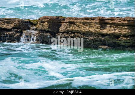 Les vagues de l'océan s'écrasent contre une formation rocheuse accidentée avec de l'eau en cascade et de l'écume marine par une journée ensoleillée. Banque D'Images