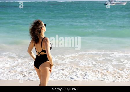 Une femme en maillot de bain noir se dresse sur une plage, face à l'eau tandis que les vagues tapent doucement sur le rivage. Elle prend dans le soleil et la brise de l'océan, profitant d'un re Banque D'Images