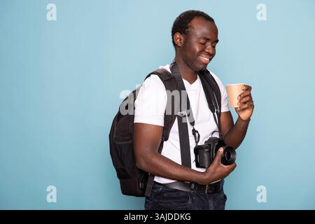 Homme afro-américain touche son objectif de caméra, souriant en regardant la tasse jetable dans sa main. Photographe masculin noir avec un sac à dos et un reflex numérique regarde son café avec une expression de contenu. Banque D'Images